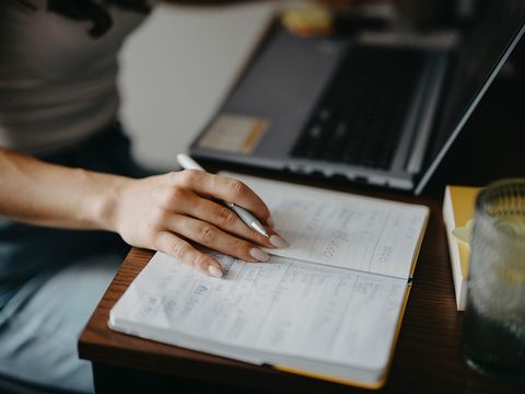 Hand mit Kugelschreiber über einem Notizbuch mit Planungsnotizen, dahinter ein aufgeklappter Laptop