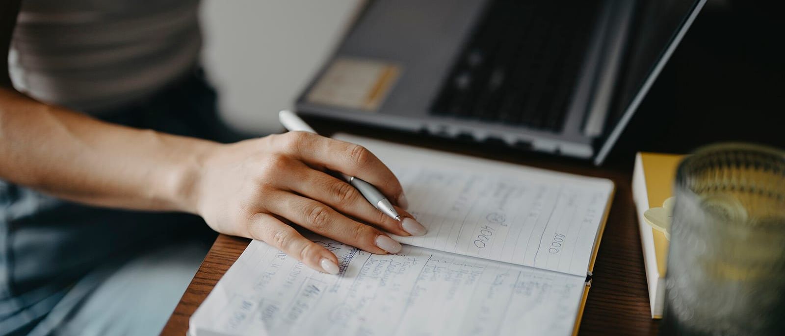 Hand mit Kugelschreiber über einem Notizbuch mit Planungsnotizen, dahinter ein aufgeklappter Laptop