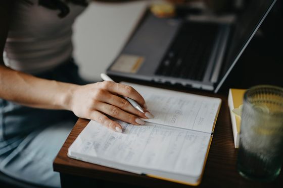Hand mit Kugelschreiber über einem Notizbuch mit Planungsnotizen, dahinter ein aufgeklappter Laptop