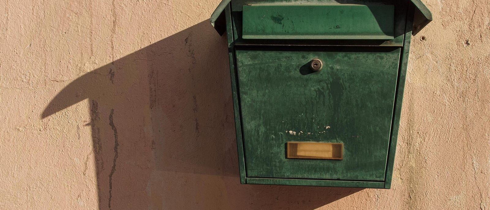 Grüner Metallbriefkasten mit Namensschild an einer sonnigen, verputzten Hauswand